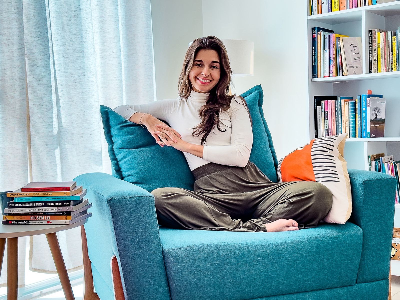 Vitória Estrela Pinto sorrindo em seu consultório, sentada em uma poltrona azul diante de uma estante de livros